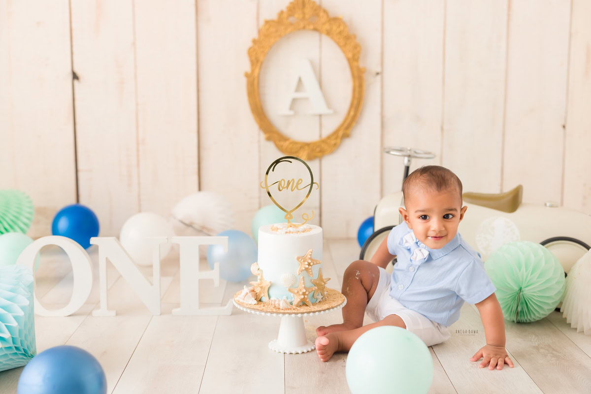 1 year old boy with Cake Smash, featuring an alphabet frame on the wall, a beige backdrop, pompom decorations, and balloons, captured in a delightful first birthday photo shoot by Anega Bawa in Delhi, Gurgaon.