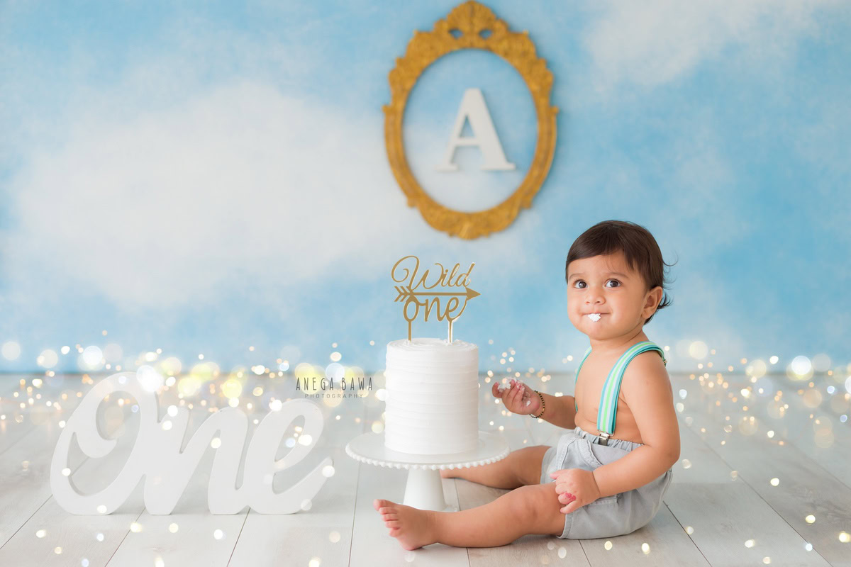 1 year boy with a cake, set against a blue backdrop featuring an alphabet frame on the wall and fairy lights during a first birthday photoshoot by well-known Anega Bawa Photography in Delhi, Gurgaon.