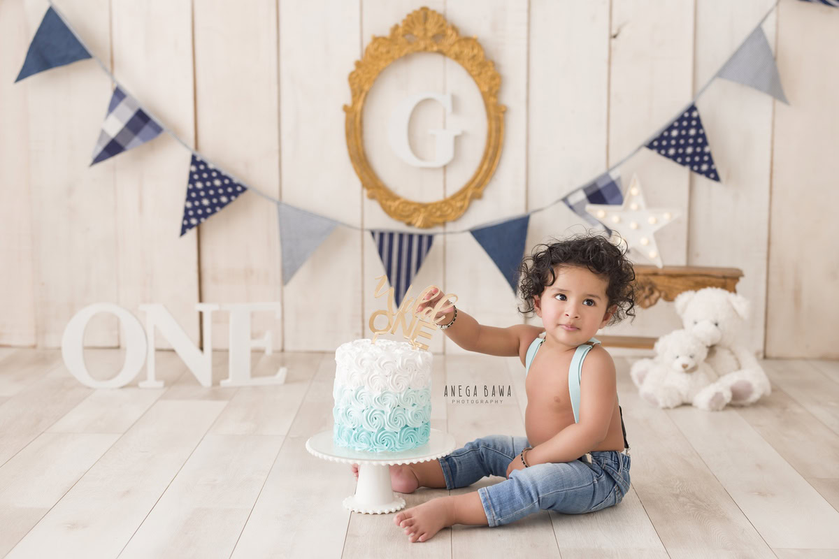 1 year old boy with Cake Smash, set against a beige backdrop, featuring an alphabet frame on the wall and a teddy bear, captured in a charming first birthday photoshoot by Anega Bawa in Delhi, Gurgaon.