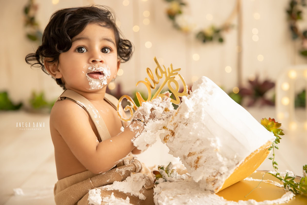 1year old boy delighting in a cake smash against a beige backdrop with enchanting fairy lights. Captured during a memorable first birthday photo shoot in Delhi, Gurgaon by the well-known family photographer Anega Bawa.