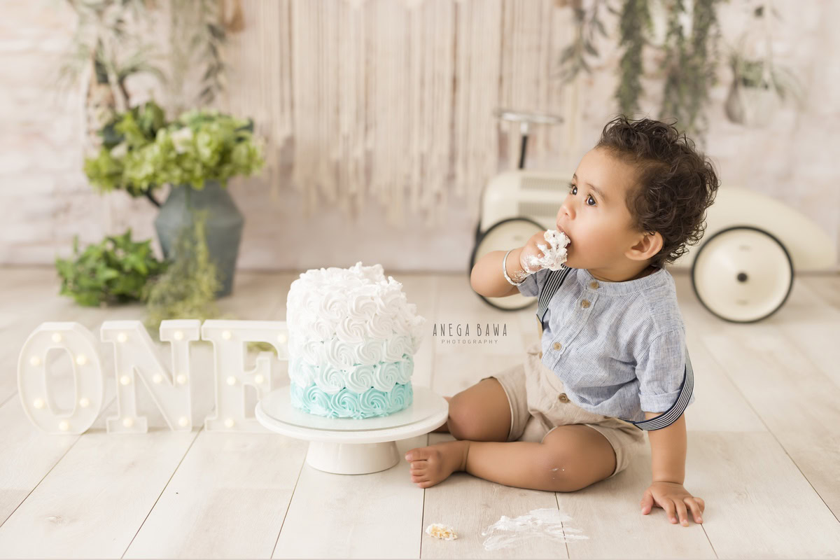 1 year boy with a cake, set against a beige backdrop with a green vase and toy scooter during a first birthday photography session by well-known Anega Bawa Photography in Delhi, Gurgaon.
