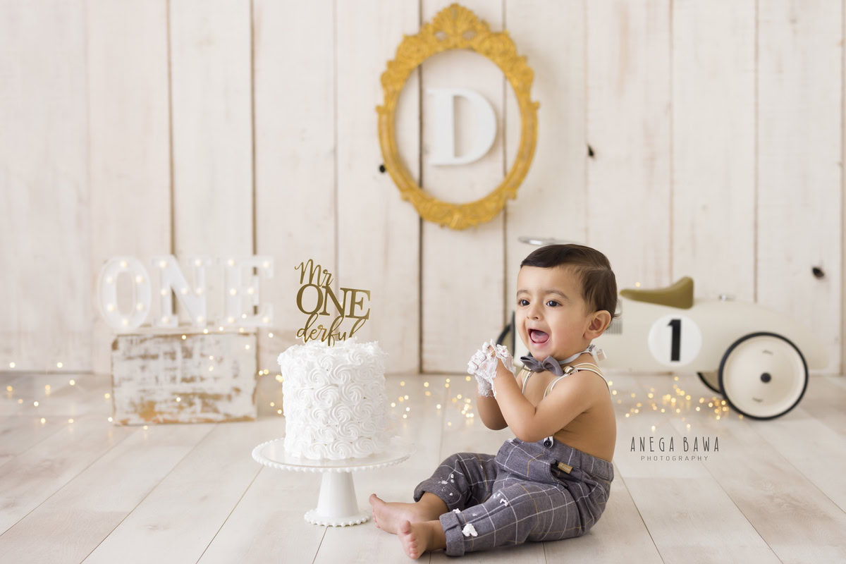 1year old boy with cake smash, beige backdrop, toy scooter, and an alphabet wooden frame on the wall. Captured during a charming first birthday photoshoot in Delhi, Gurgaon by the acclaimed family photographer Anega Bawa.