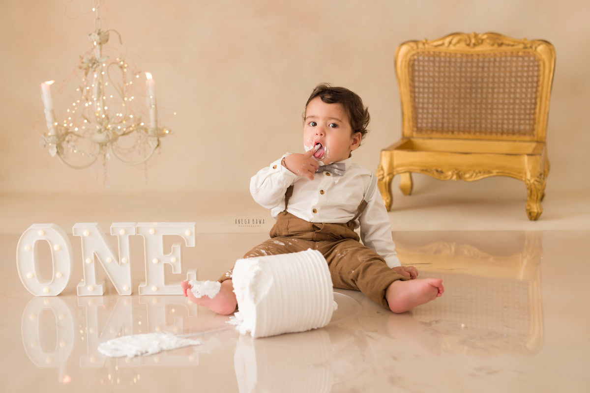 1 year boy with a cake, set against a beige backdrop featuring a wooden chair, candle stand, and fairy lights during a first birthday photoshoot by well-known Anega Bawa Photography in Delhi, Gurgaon.
