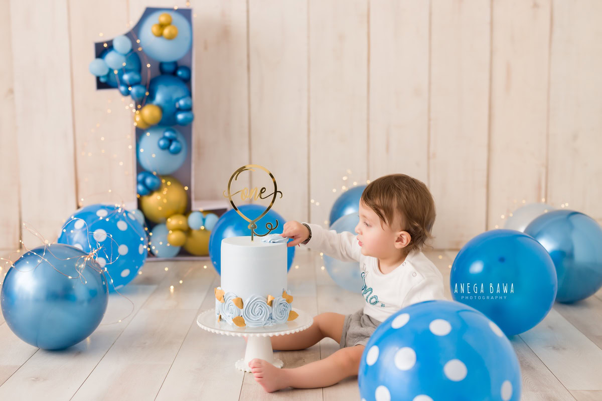 1year old boy with blue balloons, enjoying a cake smash session with a beige backdrop, celebrating his first birthday. Cake Smash photo shoot in Delhi, Gurgaon by Anega Bawa Family Photographer Gurgaon (Delhi NCR).