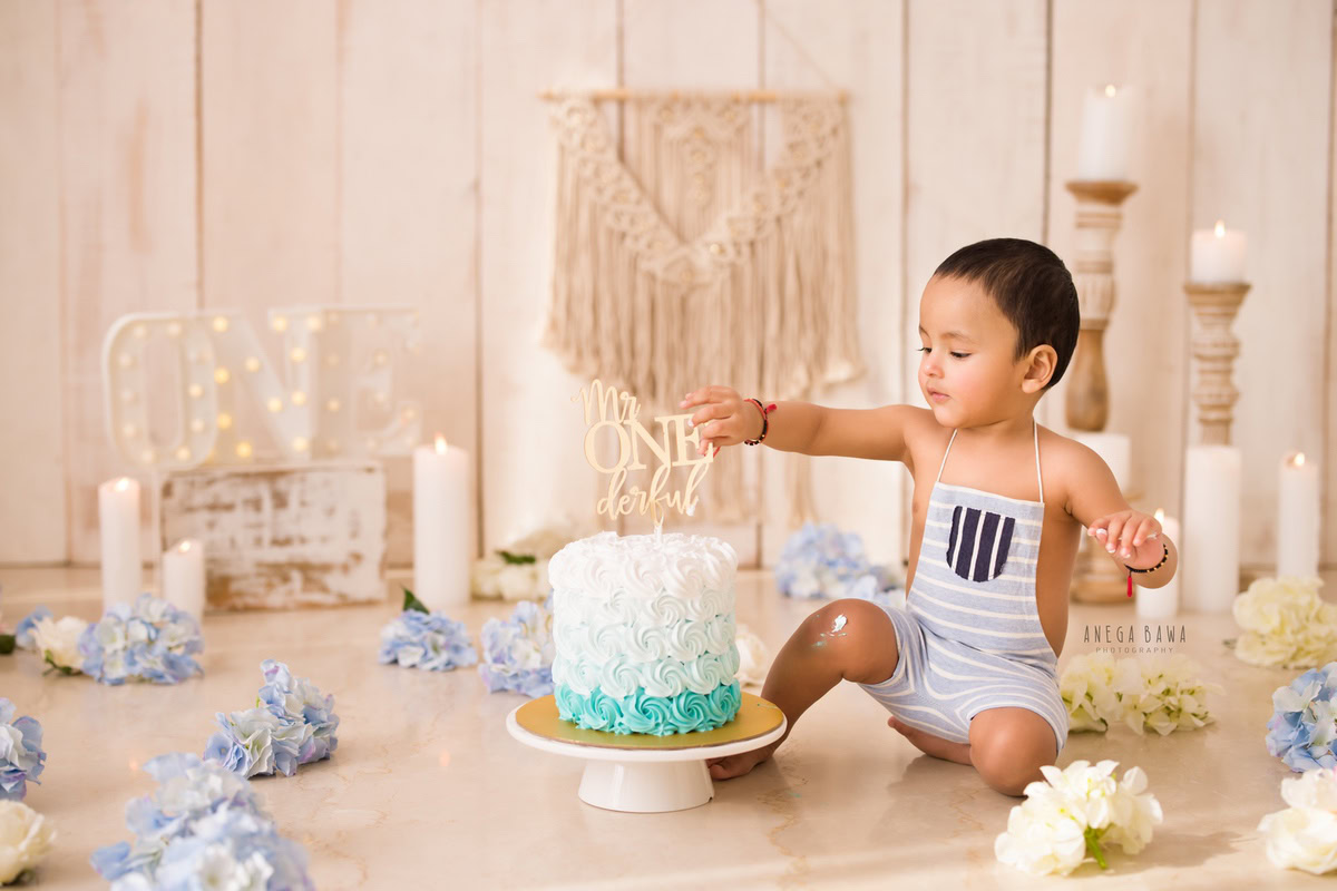 1year old boy with blue and ivory flowers, fairy lights, and candle stand with candles, enjoying a cake smash session. Celebrating his first birthday. Cake Smash photo shoot in Delhi, Gurgaon by Anega Bawa Family Photographer Gurgaon (Delhi NCR).