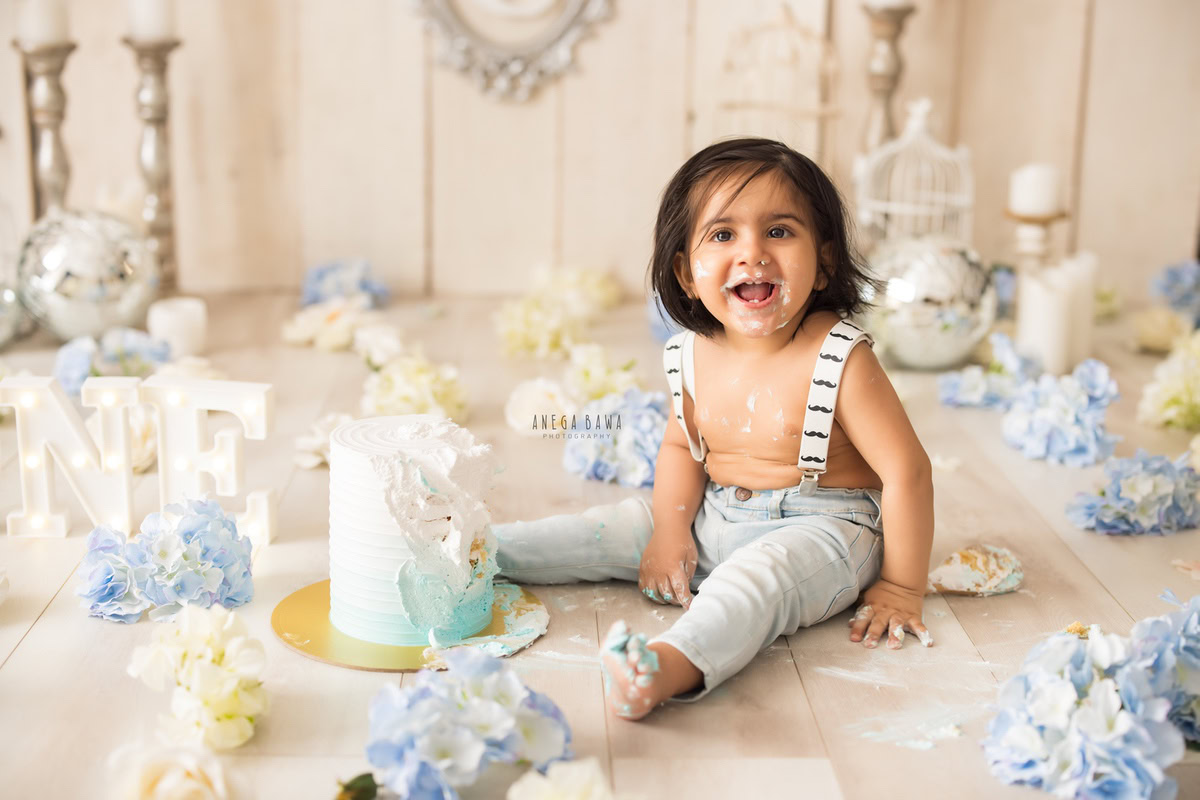 1year old boy with blue and white flowers, white castles, cake smash stand, and disco stand, enjoying a cake smash session with a beige backdrop. Celebrating his first birthday. Cake Smash photo shoot in Delhi, Gurgaon by Anega Bawa Family Photographer Gurgaon (Delhi NCR).