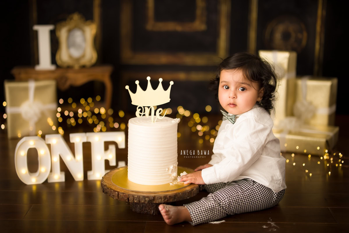 1year old boy with a brown backdrop, fairy lights, golden frame, and gifts, enjoying a cake smash session. Celebrating his first birthday. Cake Smash photo shoot in Delhi, Gurgaon by Anega Bawa Family Photographer Gurgaon (Delhi NCR).