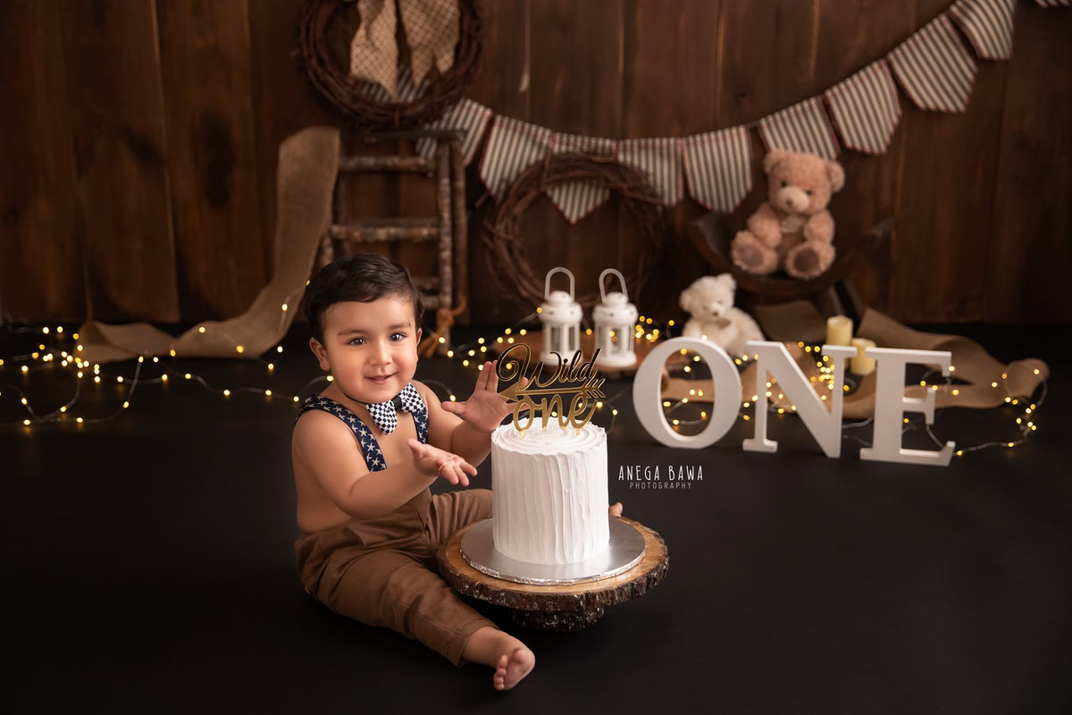 1year old boy with blue and white flowers, white castles, cake smash stand, and disco stand, enjoying a cake smash session with a beige backdrop. Celebrating his first birthday. Cake Smash photo shoot in Delhi, Gurgaon by Anega Bawa Family Photographer Gurgaon (Delhi NCR).