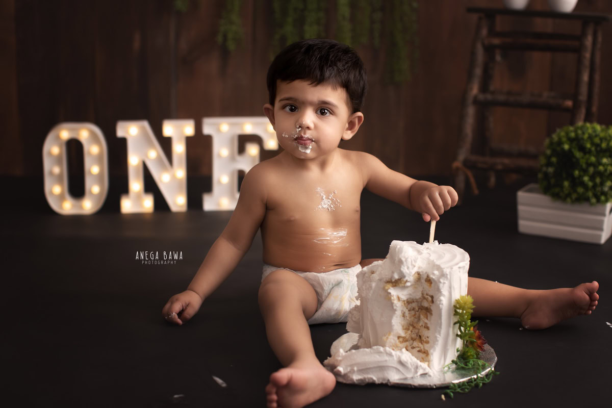 1year old boy enjoying a cake smash with a brown backdrop, white pot, and green leaves. Captured during a vibrant first birthday photoshoot in Delhi, Gurgaon by renowned family photographer Anega Bawa.