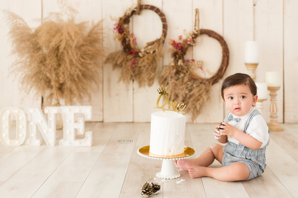 1 year old boy with Cake Smash, surrounded by bushes, a candle stand, and a round wooden frame on the wall, captured in a delightful first birthday photoshoot by Anega Bawa in Delhi, Gurgaon.