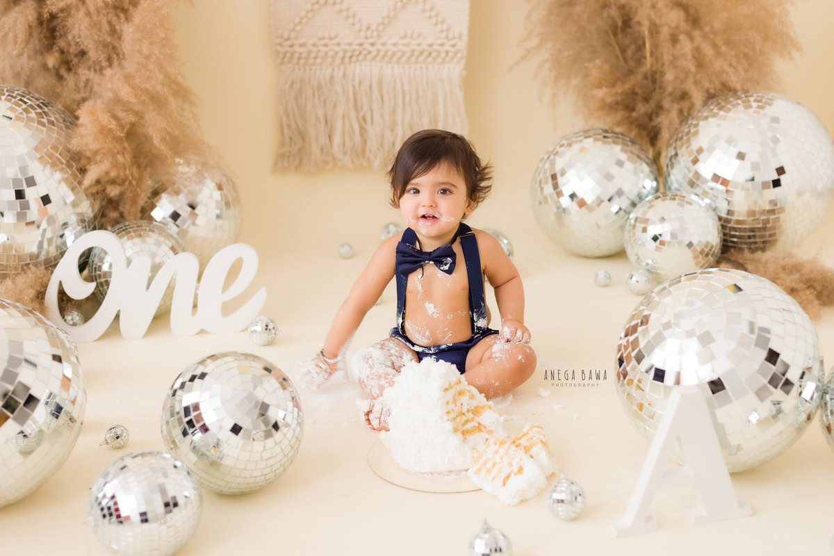 1year old boy with cake smash, surrounded by bushes and discoballs, set against a beige backdrop. Captured during an engaging first birthday photography session in Delhi, Gurgaon by the esteemed family photographer Anega Bawa.
