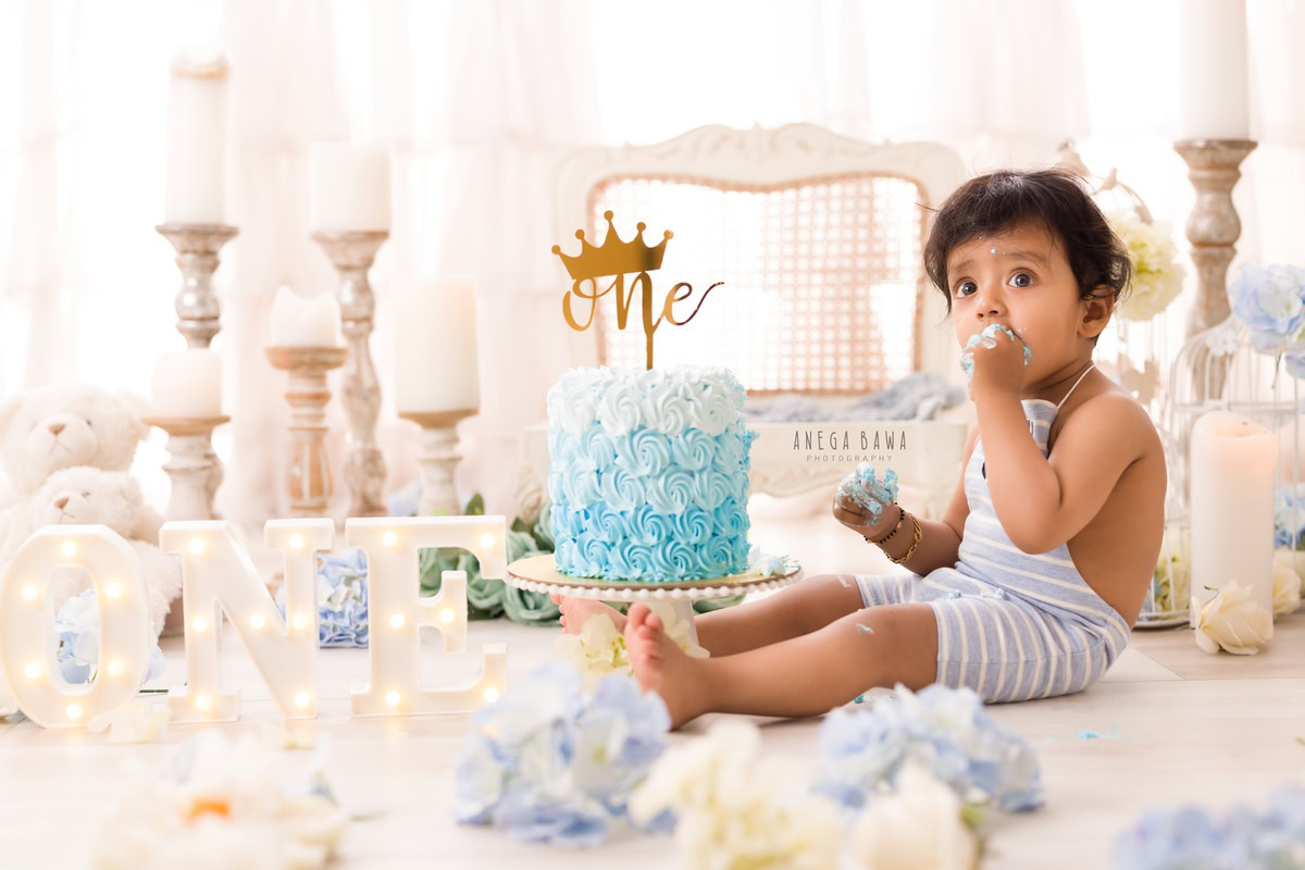 1year old boy with a candle stand, white wooden chair, blue and white flowers, enjoying a cake smash session. Celebrating his first birthday. Cake Smash photo shoot in Delhi, Gurgaon by Anega Bawa Family Photographer Gurgaon (Delhi NCR).