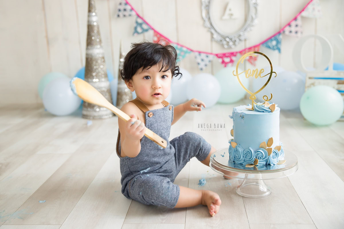 1 year boy with a cake, set against a beige backdrop featuring colorful fringe on the wall and balloons during a first birthday photoshoot by well-known Anega Bawa Photography in Delhi, Gurgaon.