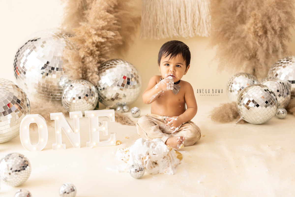 1 year old boy Cake Smash first birthday photography in Delhi, Gurgaon with disco balls, bushes, and beige backdrop. Captured by Anega Bawa family photographer Gurgaon (Delhi NCR)