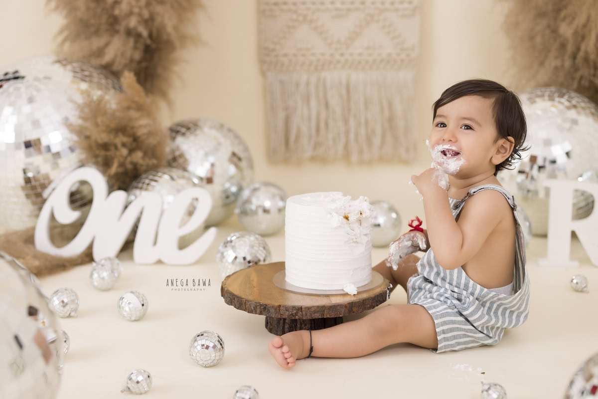 1 year old boy with Cake Smash, featuring discoballs, bushes, and a net on a beige backdrop, captured in a delightful first birthday photoshoot by Anega Bawa in Delhi, Gurgaon.