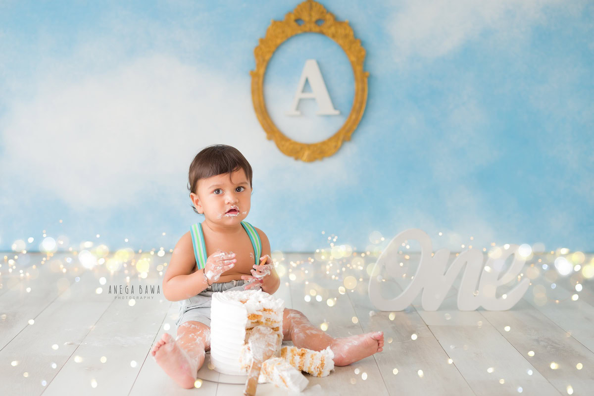 1year old boy with cake smash, surrounded by fairy lights and a blue and white backdrop. Captured during a magical first birthday photoshoot in Delhi, Gurgaon by the renowned family photographer Anega Bawa.