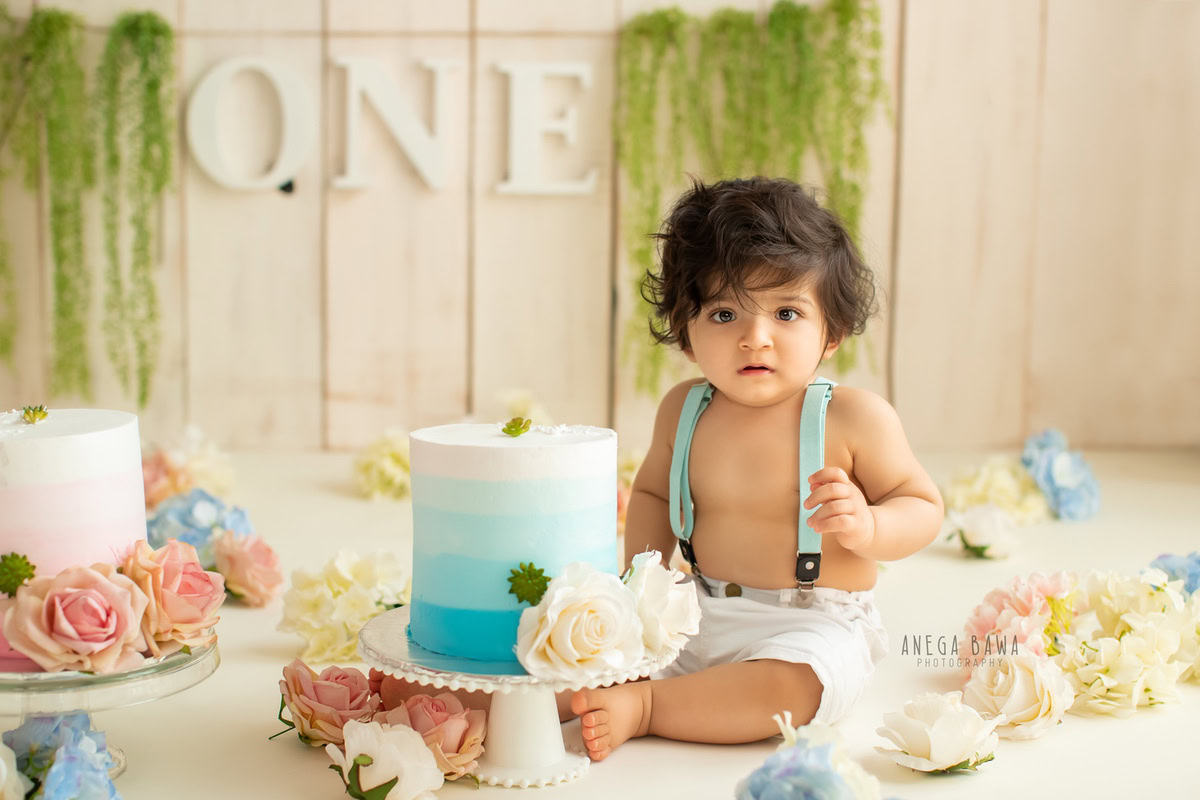 1year old boy with cake smash, leafy fringe on the wall, and flowers on the floor, set against a beige backdrop. Captured during a delightful first birthday photography session in Delhi, Gurgaon by the renowned family photographer Anega Bawa.