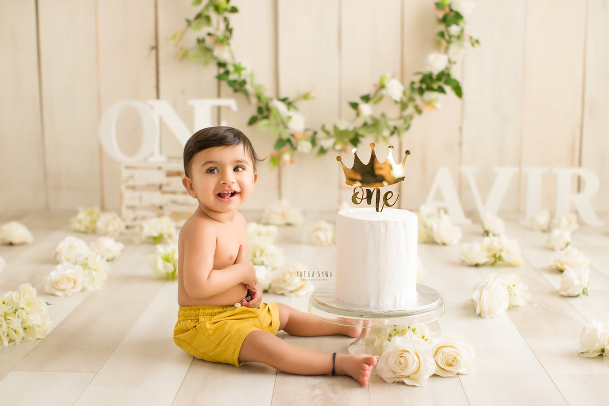 1year old boy with a name frame on the floor, white flowers, floral fringe on the wall, and a beige backdrop, enjoying a cake smash session. Celebrating his first birthday. Cake Smash photo shoot in Delhi, Gurgaon by Anega Bawa Family Photographer Gurgaon (Delhi NCR).