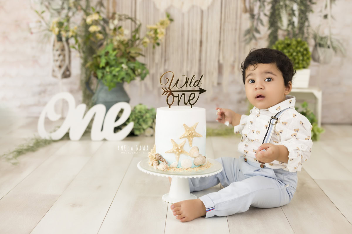 1 year old boy with Cake Smash, set against a net beige backdrop, with a green vase and flowers, captured in a charming first birthday photoshoot by Anega Bawa in Delhi, Gurgaon.