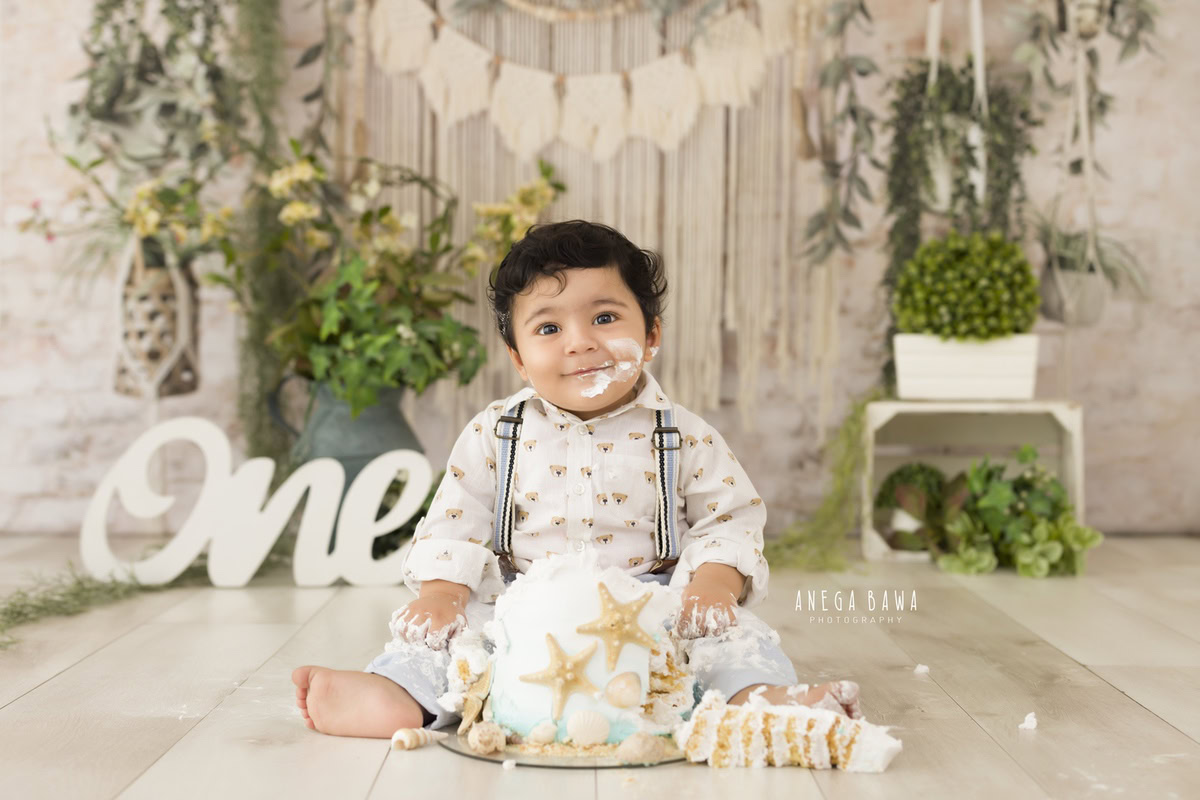 1 year old boy with Cake Smash, against a net beige backdrop, featuring a green vase and leafy fringes, captured in a charming first birthday photography session by Anega Bawa in Delhi, Gurgaon.