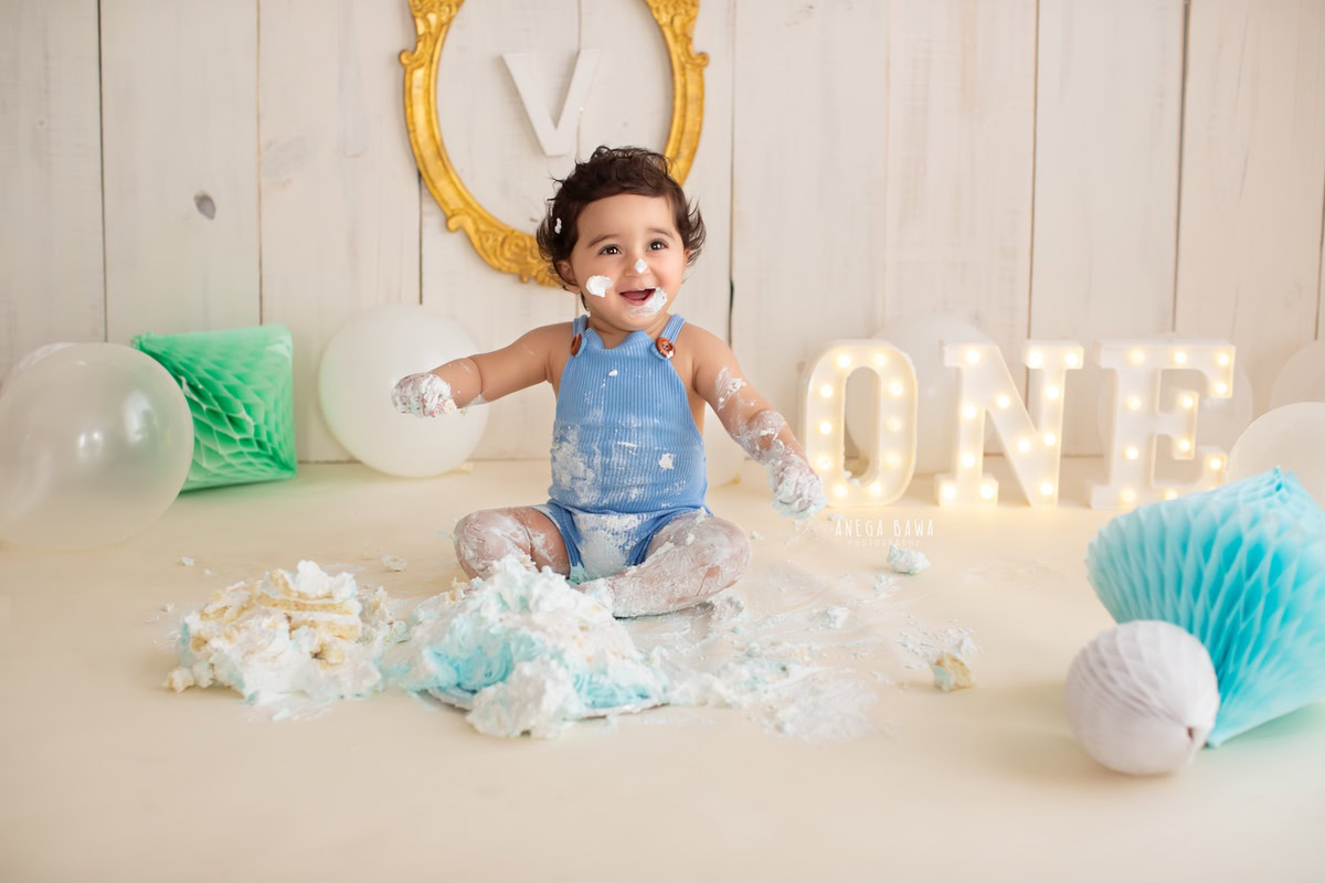 1 year old boy Cake Smash on face first birthday photoshoot in Delhi, Gurgaon with beige backdrop, pompom alphabet, and golden frame on wall. Captured by Anega Bawa family photographer Gurgaon (Delhi NCR)