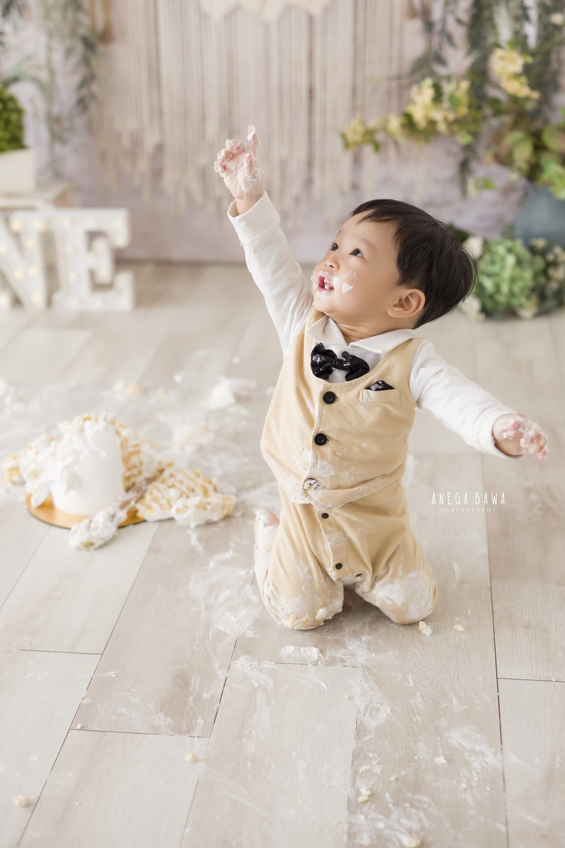 1 year old boy with Cake Smash on face, against a beige net backdrop, captured in a delightful first birthday photo shoot by Anega Bawa in Delhi, Gurgaon.
