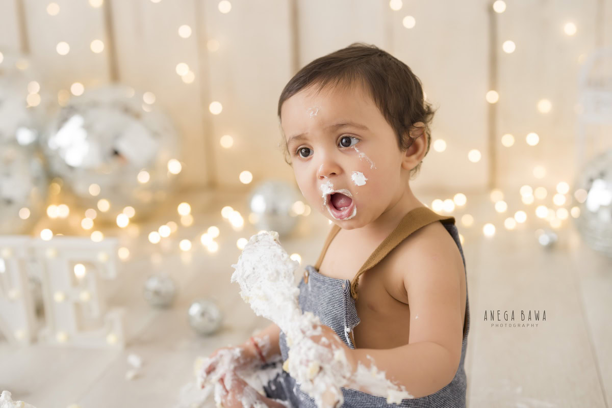 1 year old boy Cake Smash on face first birthday photo shoot in Delhi, Gurgaon with disco balls, fairy lights, and beige backdrop. Captured by Anega Bawa family photographer Gurgaon (Delhi NCR)