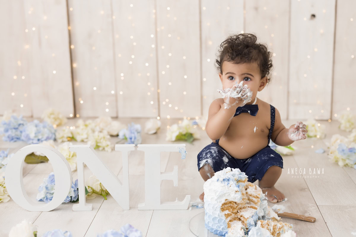 1 year old boy Cake Smash on face first birthday photoshoot in Delhi, Gurgaon with fairy lights, beige backdrop, blue and white flowers. Captured by Anega Bawa family photographer Gurgaon (Delhi NCR)
