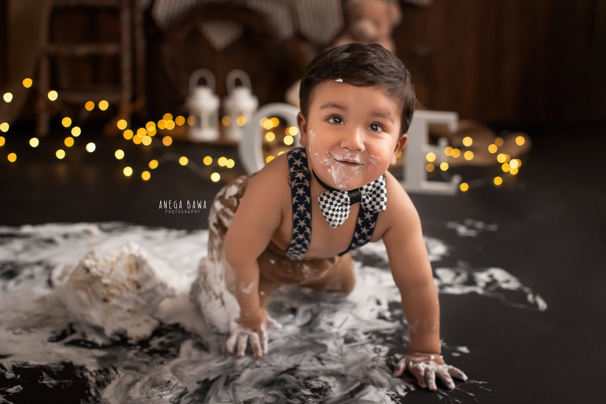 1 year boy with cake on his face and hands, set against a brown backdrop with fairy lights during a first birthday photoshoot by well-known Anega Bawa Photography in Delhi, Gurgaon.