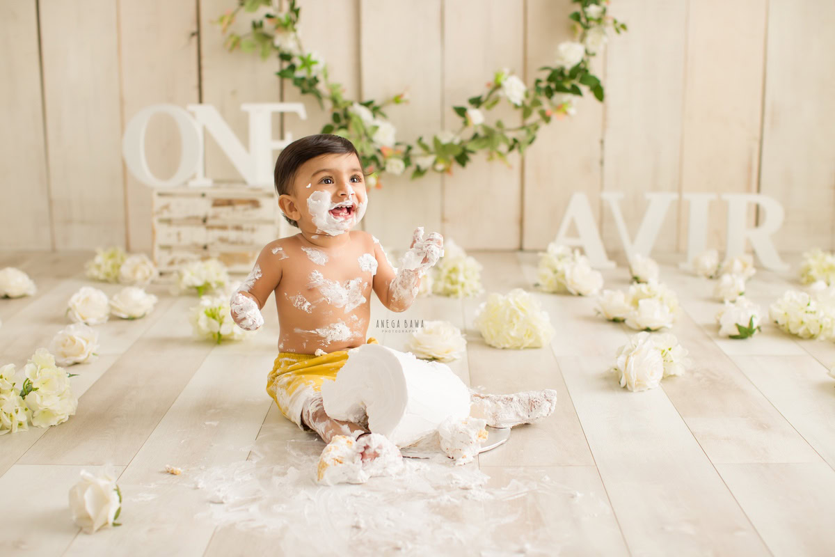 1 year old boy Cake Smash on face first birthday photography in Delhi, Gurgaon with white flowers, name frame on floor, and beige backdrop. Captured by Anega Bawa family photographer Gurgaon (Delhi NCR)