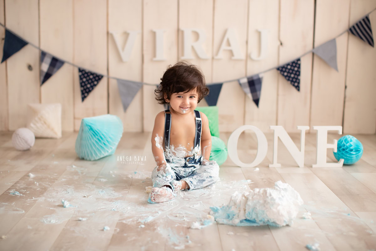 1year old boy with cake smash pompom decorations beige backdrop and fringe on the wall. Captured during a delightful first birthday photography session in Delhi Gurgaon by the renowned family photographer Anega Bawa.