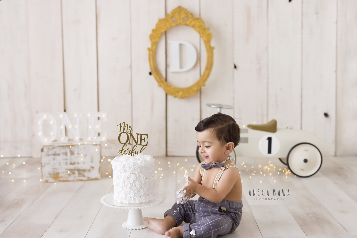 1 year old boy with Cake Smash, featuring a toy scooter, beige backdrop, fairy lights, and an alphabet frame on the wall, captured in a delightful first birthday photoshoot by Anega Bawa in Delhi, Gurgaon.
