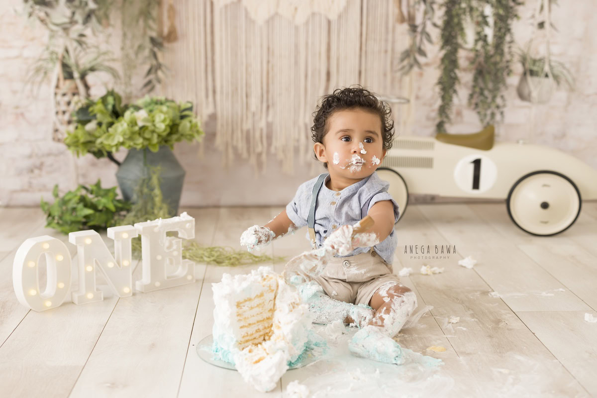 1 year old boy with Cake Smash, featuring a toy scooter, beige backdrop, and a green flower vase, captured in a charming first birthday photography session by Anega Bawa in Delhi, Gurgaon.