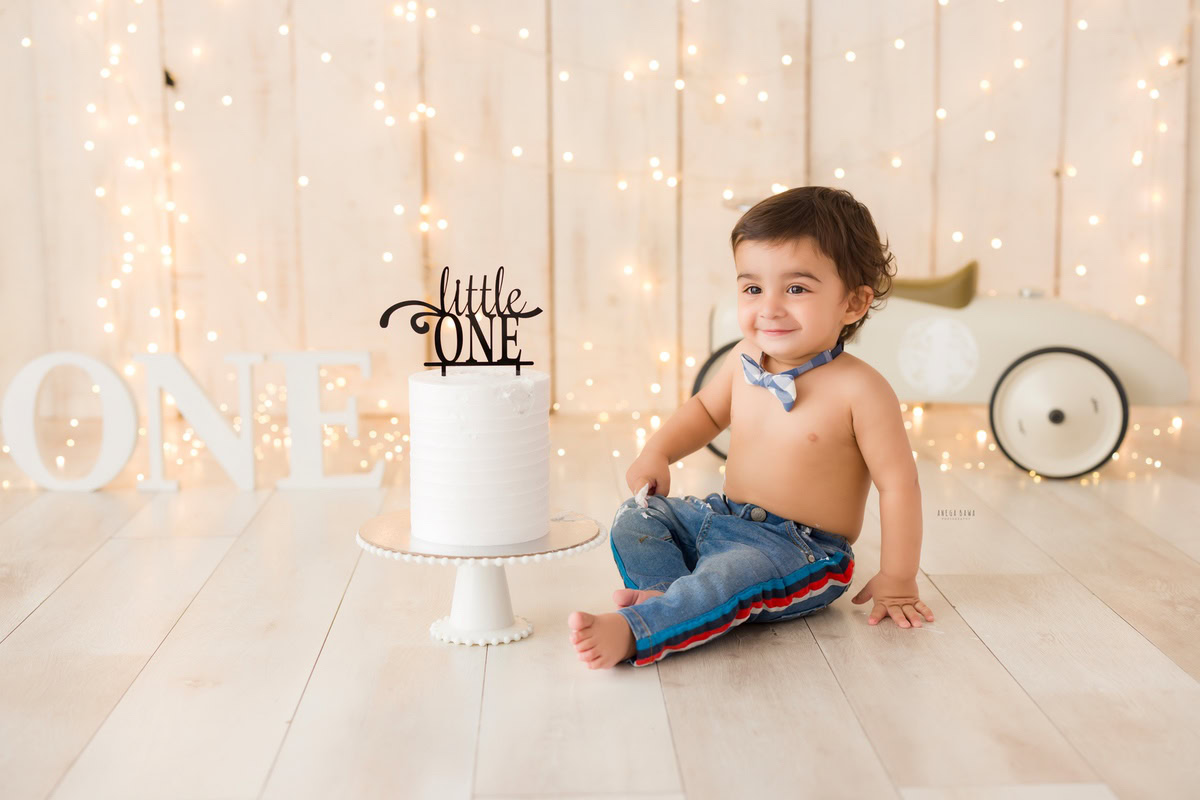 1 year old boy with Cake Smash, featuring a toy scooter and fairy lights against a beige backdrop, captured in a delightful first birthday photo shoot by Anega Bawa in Delhi, Gurgaon.