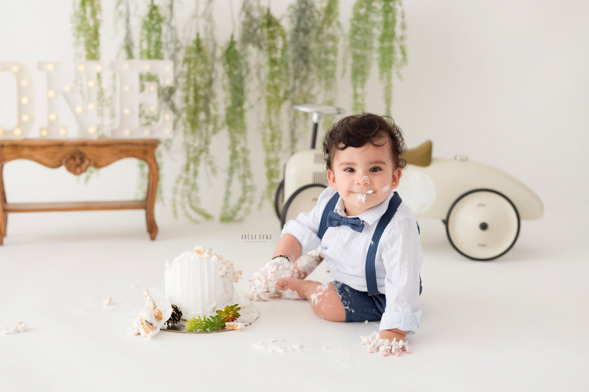 1 year boy with a cake, surrounded by a toy scooter and leafy fringes on the wall against a white backdrop during a first birthday photoshoot by well-known Anega Bawa Photography in Delhi, Gurgaon.
