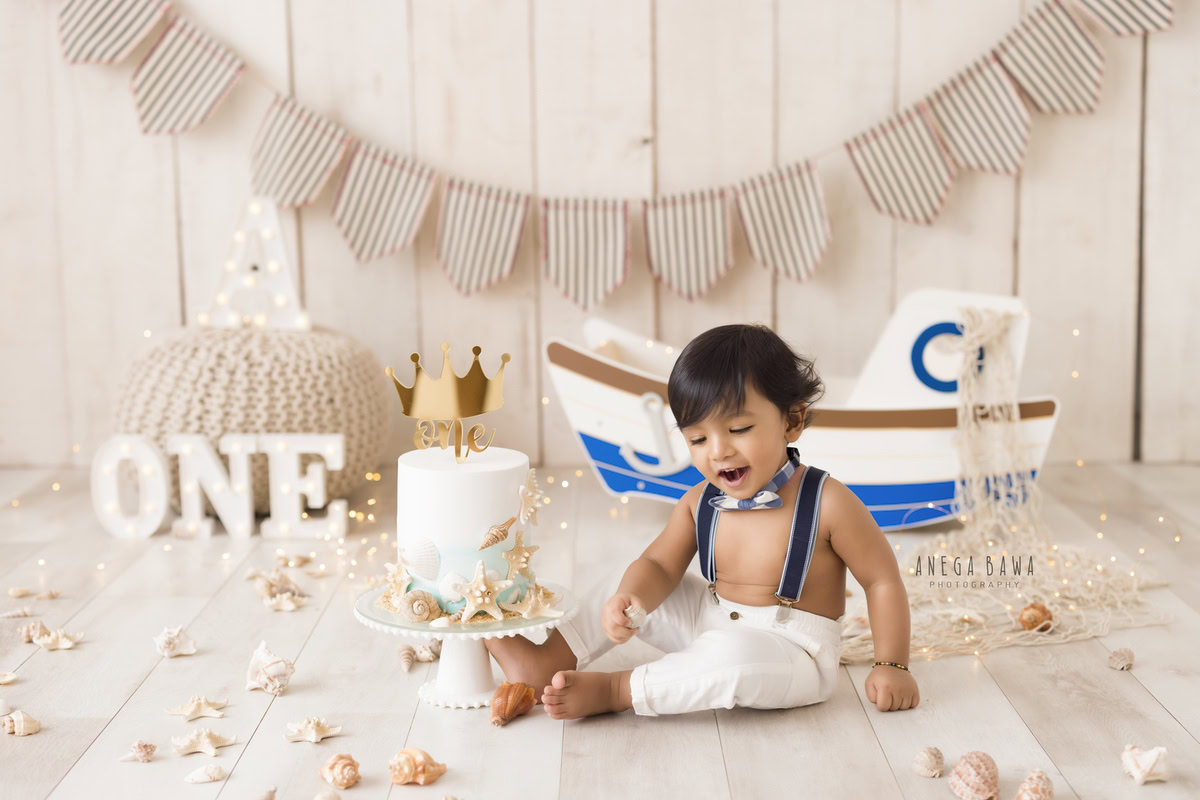 1 year old boy with Cake Smash, featuring a toy spaceship and flowers on the floor against a beige backdrop, captured in a charming first birthday photoshoot by Anega Bawa in Delhi, Gurgaon.