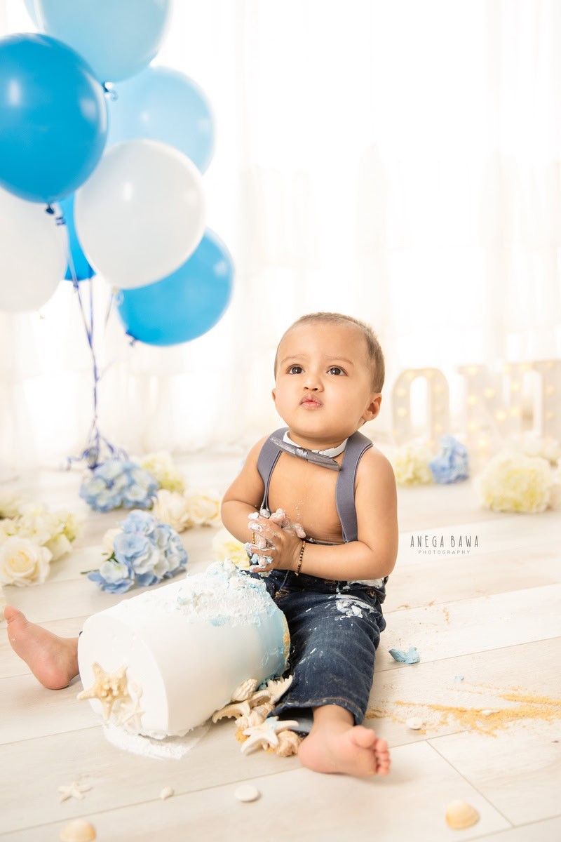 1 year boy with a cake, surrounded by white and blue balloons against a white backdrop during a first birthday photoshoot by well-known Anega Bawa Photography in Delhi, Gurgaon.