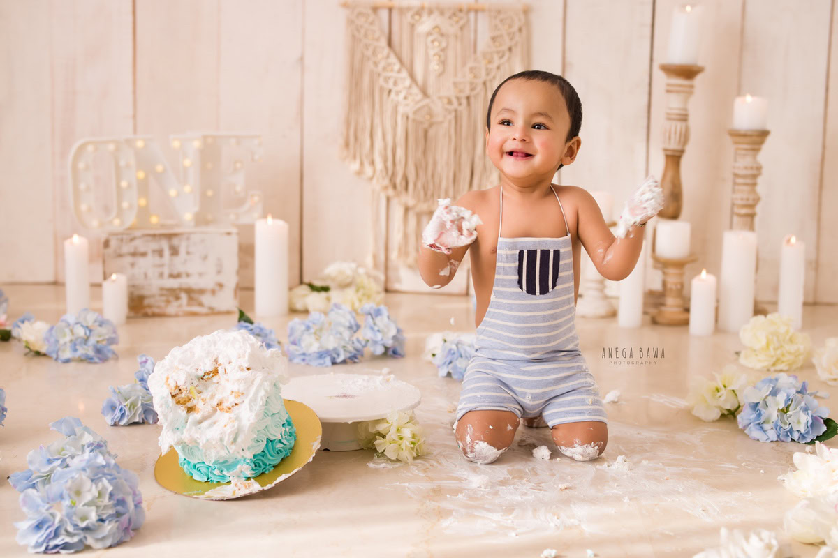 1 year old boy Cake Smash first birthday photoshoot in Delhi, Gurgaon with white candles, beige backdrop, and blue flowers. Captured by Anega Bawa family photographer Gurgaon (Delhi NCR)