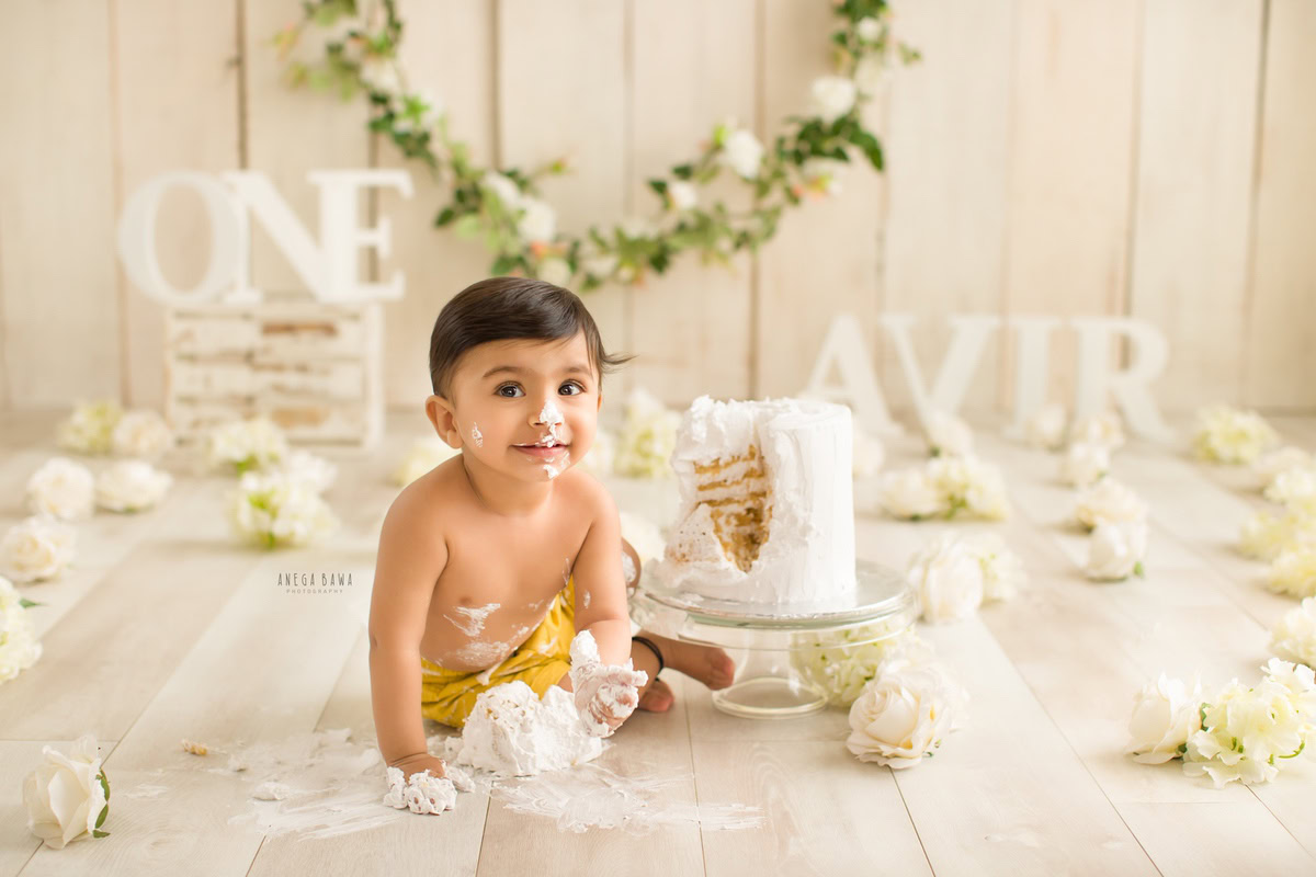 1 year boy with a cake, surrounded by white flowers and set against a beige backdrop with a name frame on the wall during a first birthday photoshoot by well-known Anega Bawa Photography in Delhi, Gurgaon.
