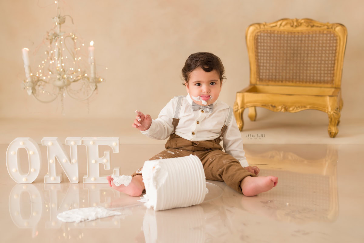 1 year boy with a cake, set against a beige backdrop featuring a wooden chair, candle stand, and fairy lights during a first birthday photoshoot by well-known Anega Bawa Photography in Delhi, Gurgaon.