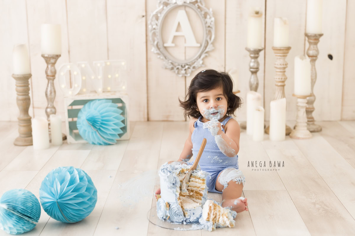 1year old boy with cake smash candle stand blue pompom beige backdrop and an alphabet frame on the wall. Captured during a charming first birthday photoshoot in Delhi Gurgaon by the acclaimed family photographer Anega Bawa.