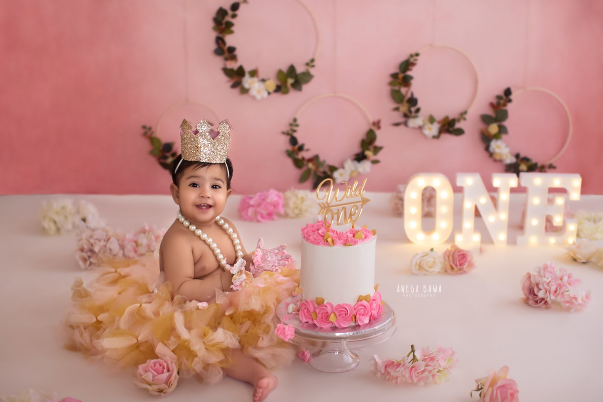 1year old boy with a crown and pearls, enjoying a cake smash session with a pink backdrop, flowers, and a wooden floral frame on the wall. Celebrating his first birthday. Cake Smash photo shoot in Delhi, Gurgaon by Anega Bawa Family Photographer Gurgaon (Delhi NCR).