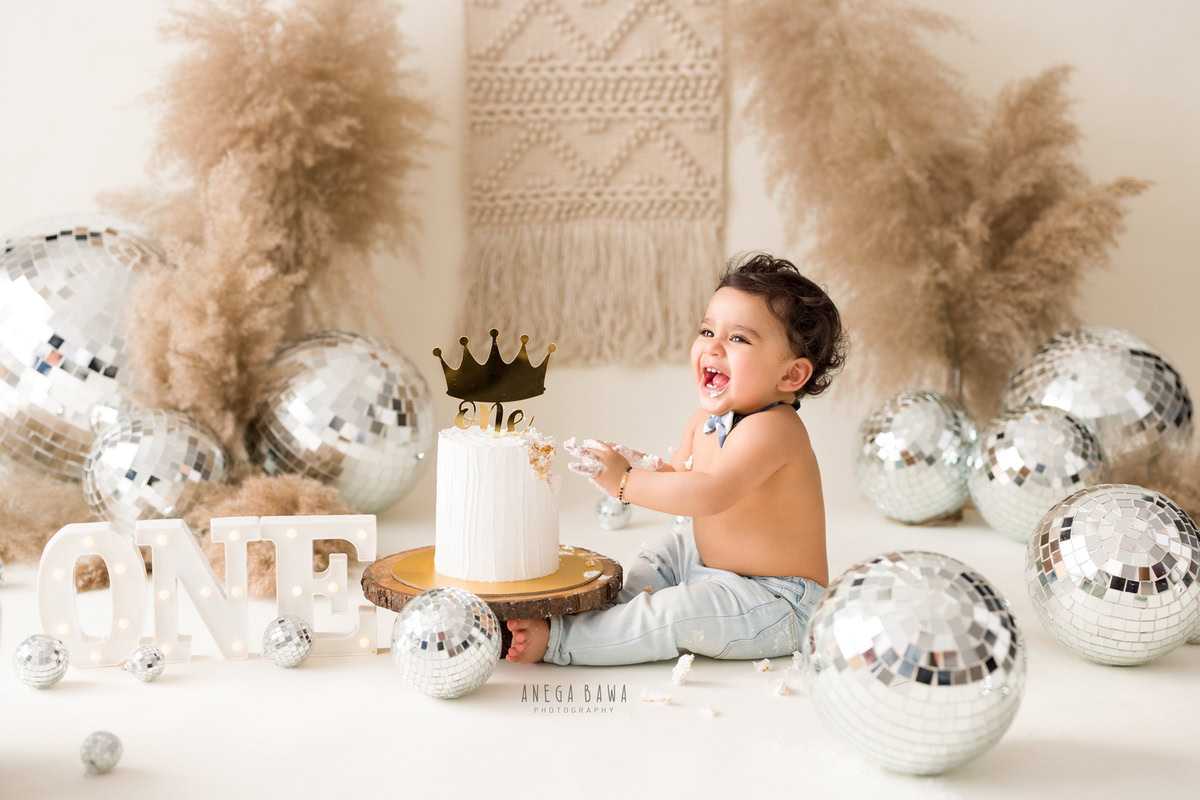 1 year old boy with Cake Smash, wearing a cute bow and surrounded by bushes and discoballs against a beige backdrop, captured in charming first birthday photography by Anega Bawa in Delhi, Gurgaon.