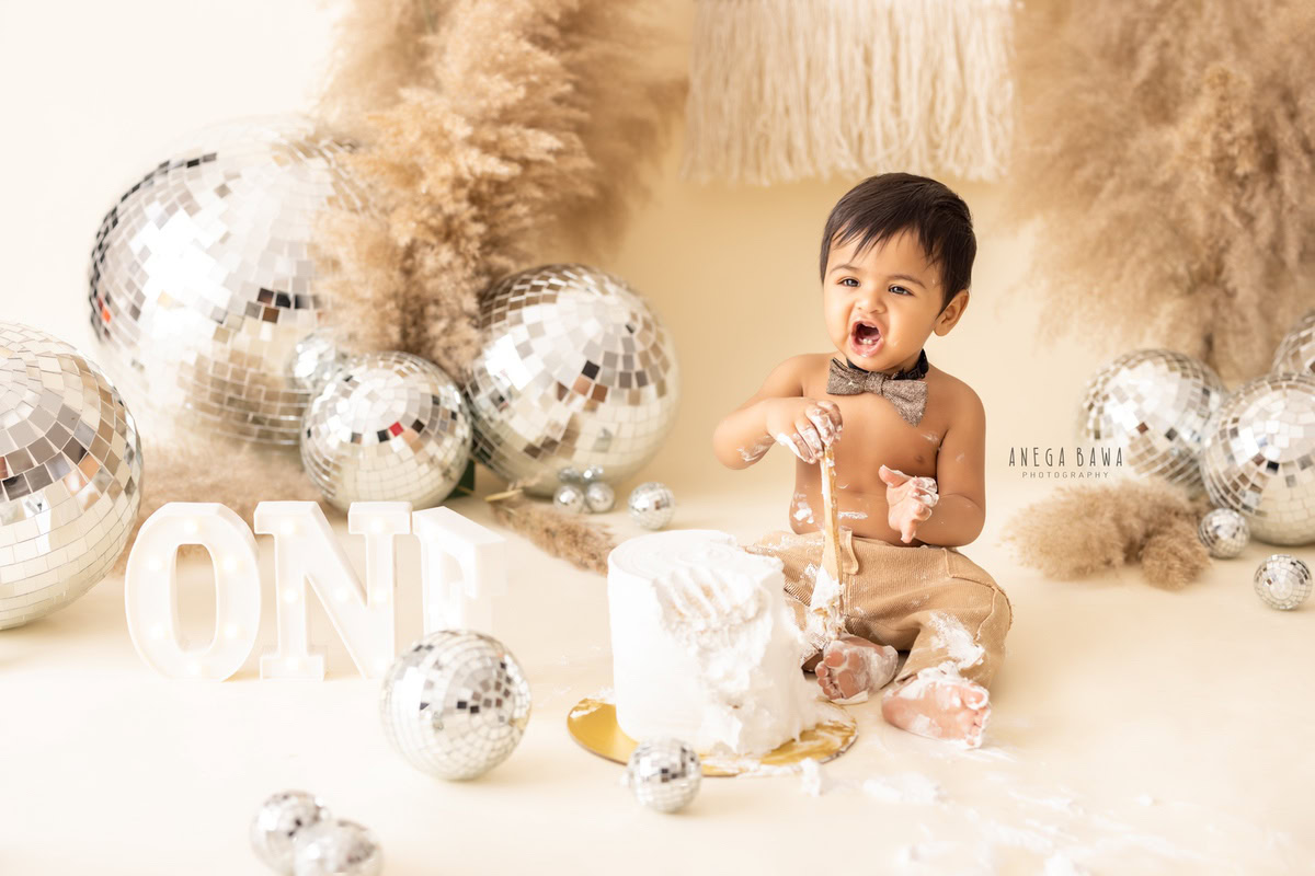 1 year old boy with Cake Smash, wearing a cute bow and playing with the cake, surrounded by discoballs and bushes, captured in a delightful first birthday photoshoot by Anega Bawa in Delhi, Gurgaon.