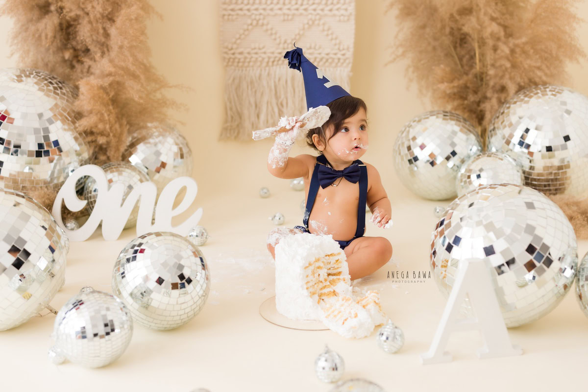1 year old boy with Cake Smash, wearing a cute cap and surrounded by bushes and discoballs against a beige backdrop, captured in a charming first birthday photoshoot by Anega Bawa in Delhi, Gurgaon.