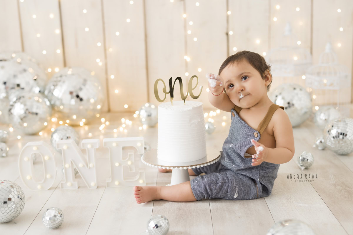 1 year boy in denim dungarees with a cake, surrounded by disco balls and fairy lights against a beige backdrop during a first birthday photoshoot by well-known Anega Bawa Photography in Delhi, Gurgaon.