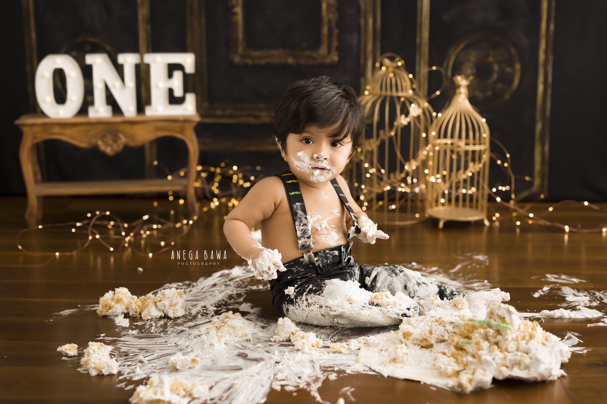 1year old boy in denim dungaree with cake smash golden castles brown backdrop and fairy lights. Captured during a delightful first birthday photo shoot in Delhi Gurgaon by the esteemed family photographer Anega Bawa.
