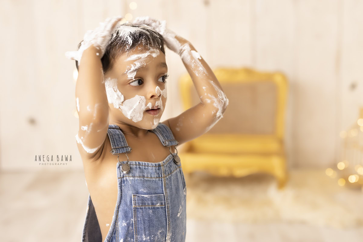 1year old boy in denim dungaree with cake smash on his face golden chair and beige backdrop. Captured during a charming first birthday photography session in Delhi Gurgaon by the renowned family photographer Anega Bawa.