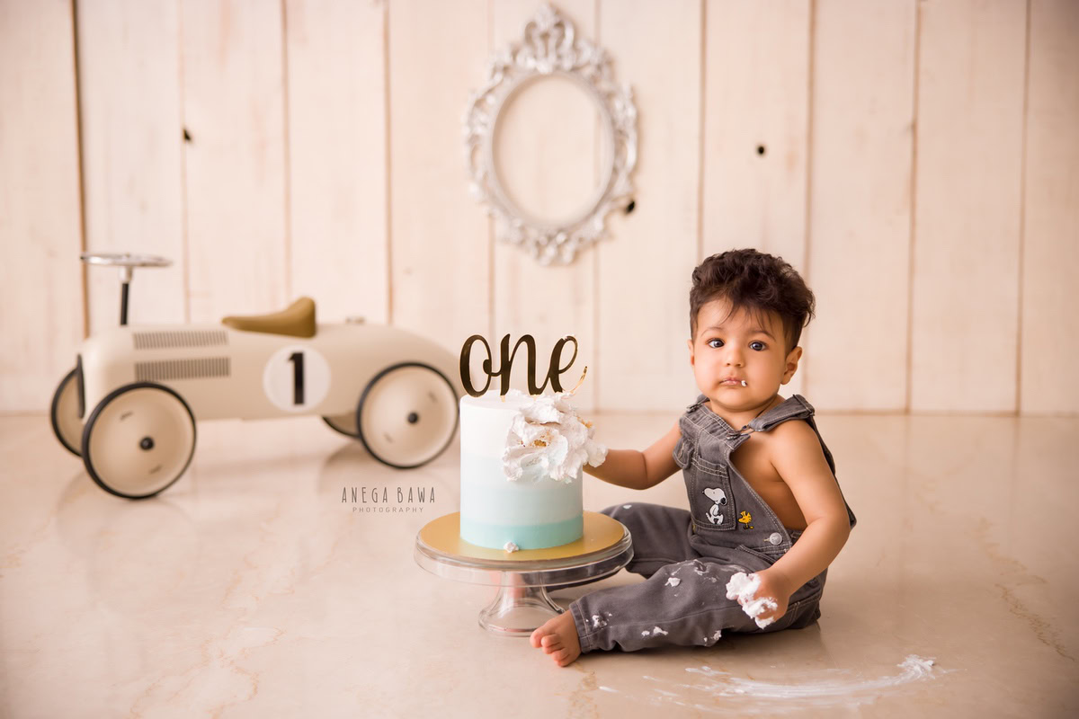 1 year old boy in denim dungaree with toy scooter beige backdrop Cake Smash first birthday photoshoot in Delhi Gurgaon. Captured by Anega Bawa family photographer Gurgaon (Delhi NCR)