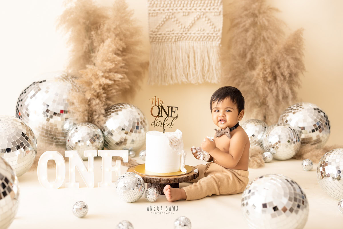 1year old boy with disco balls and bushes, enjoying a cake smash session with a beige backdrop. Celebrating his first birthday. Cake Smash photo shoot in Delhi, Gurgaon by Anega Bawa Family Photographer Gurgaon (Delhi NCR).
